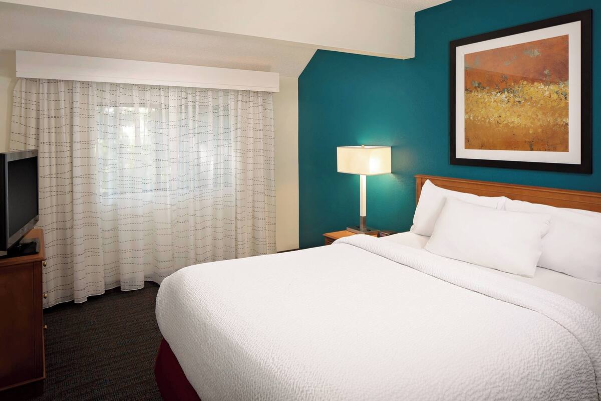 A hotel room with a large bed, a TV on a wooden cabinet, a white curtain, a teal wall, and a framed painting.