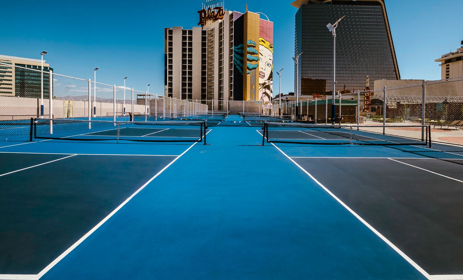 Una cancha de tenis al aire libre con una red, rodeada por una valla, y edificios al fondo.