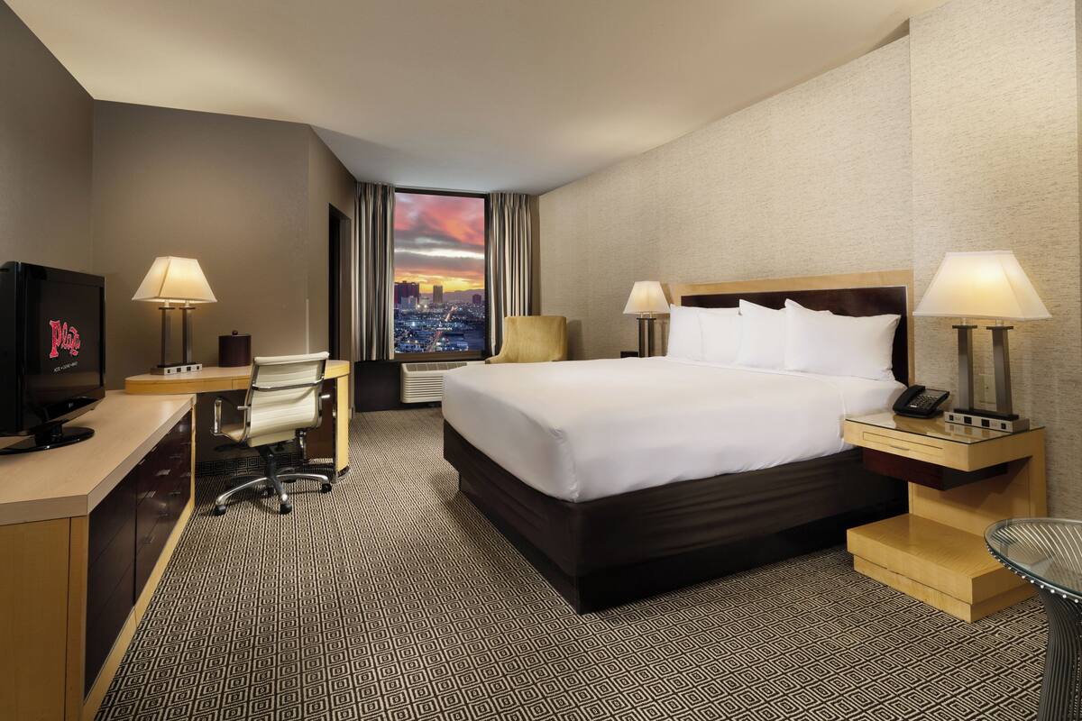 A hotel room with a large bed, two bedside tables with lamps, a desk with a chair, a TV, and a view of a cityscape at sunset.