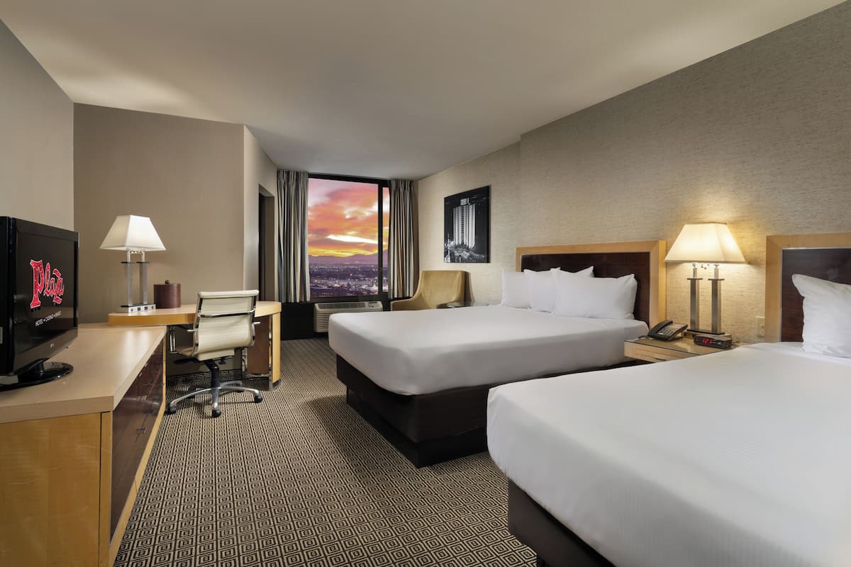 A hotel room with two beds, a desk with a computer, a TV, and a large window with a city view.
