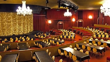A theater with rows of tables and chairs, a stage with curtains, and chandeliers.