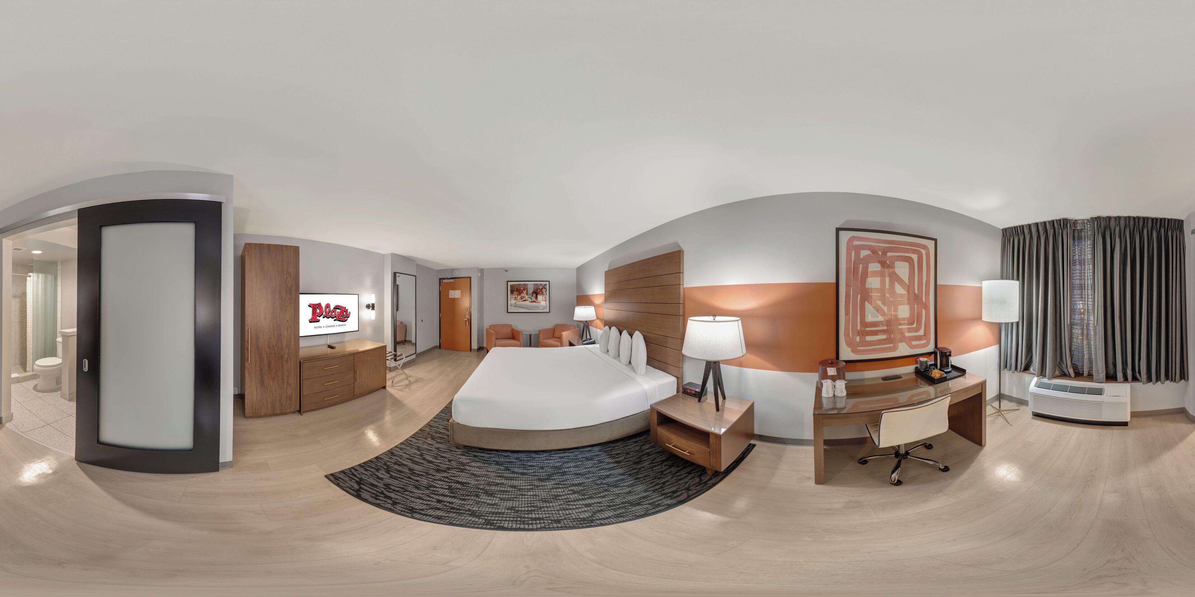 Une chambre d’hôtel moderne équipée d’un lit, d’un bureau, d’une chaise, d’une télévision et d’une salle de bain visible à travers une porte ouverte.