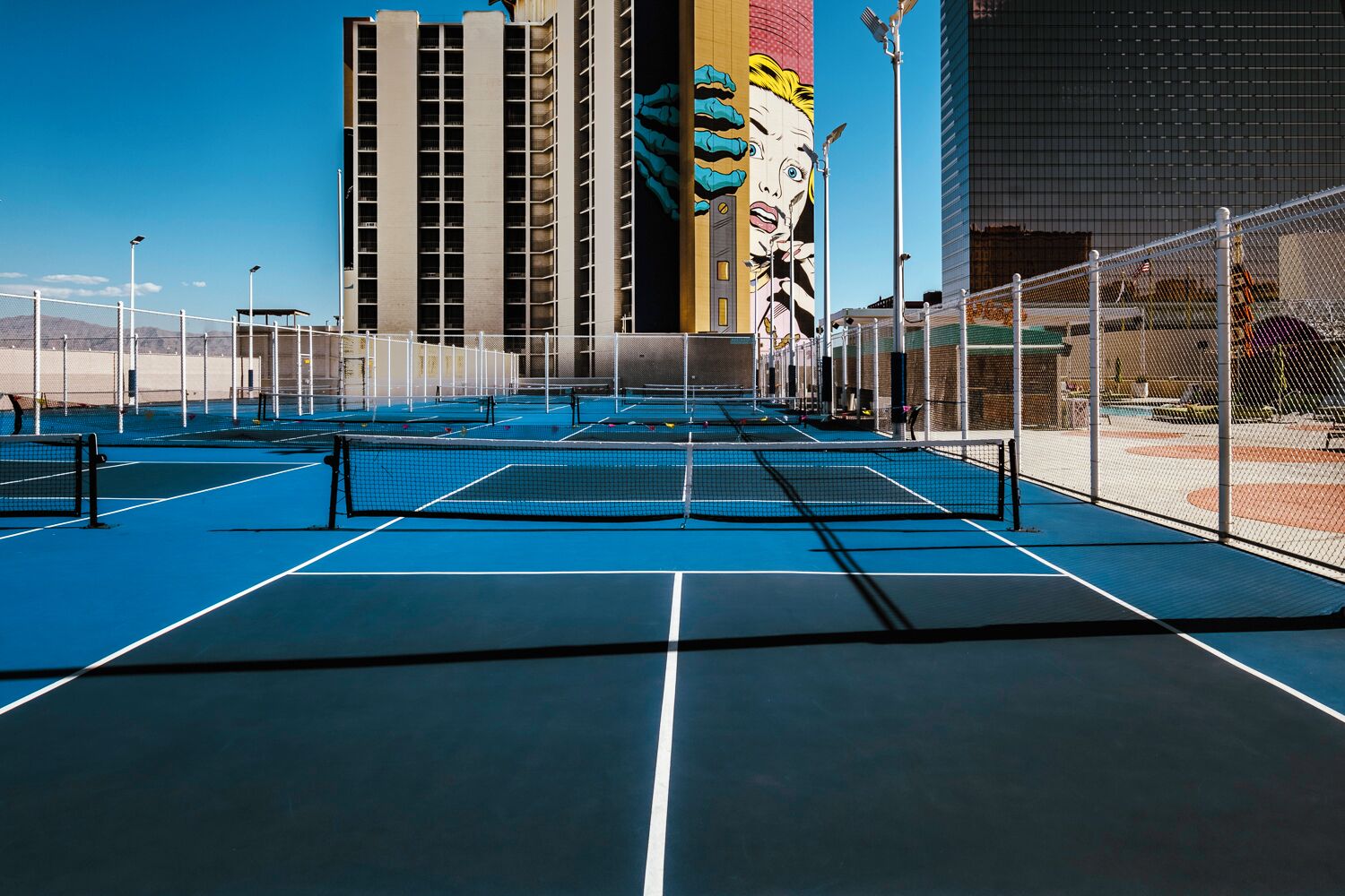 A tennis court with a net, surrounded by a fence, and buildings in the background.