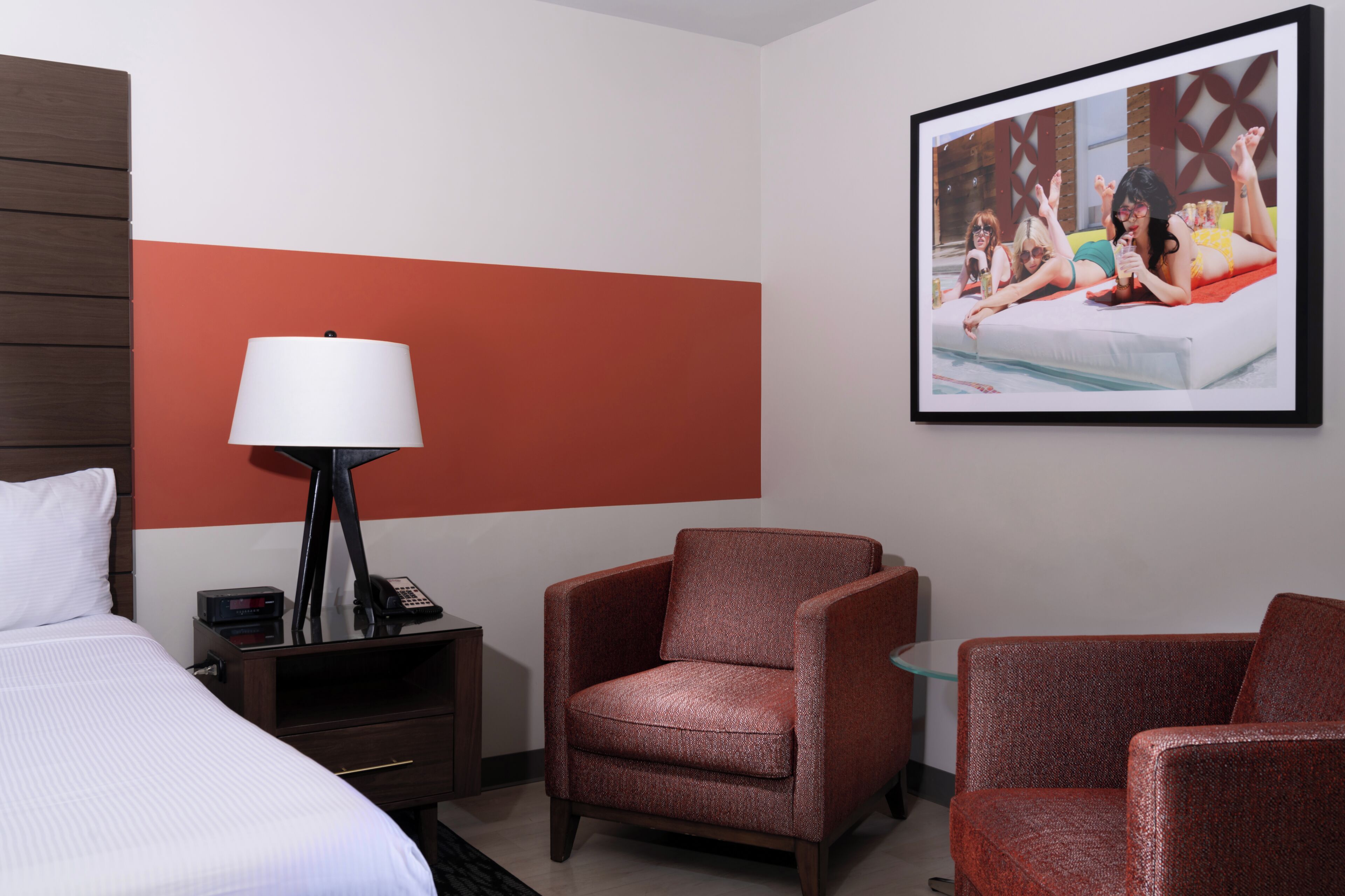 A hotel room with a bed, a nightstand with a lamp, a framed picture on the wall, and two armchairs.