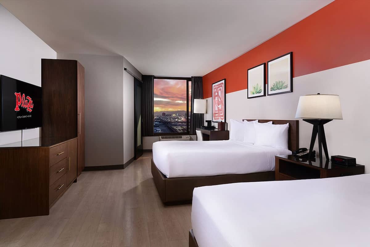 A hotel room with two beds, a TV, a desk, and a city view.
