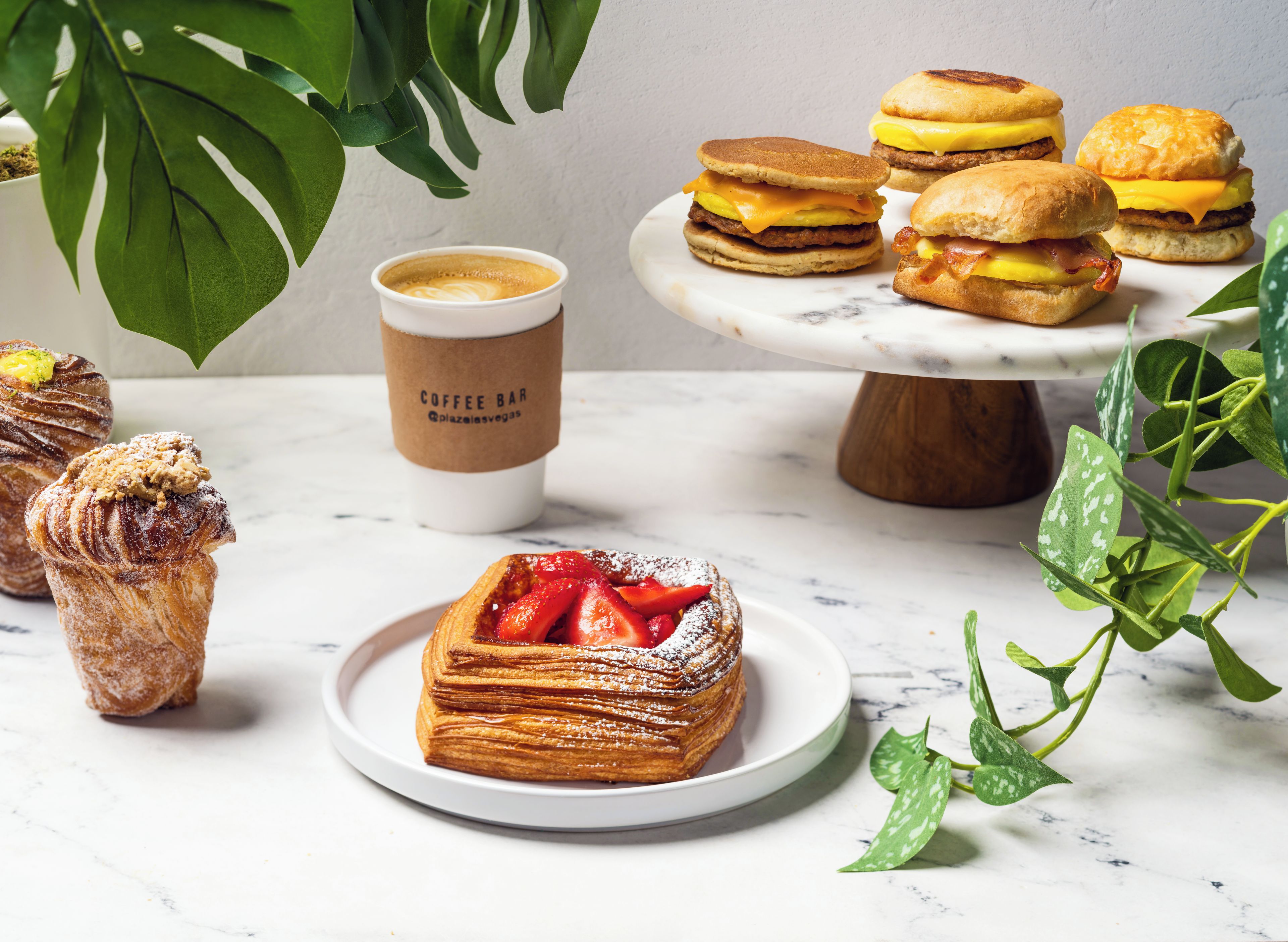 Une variété de viennoiseries et de sandwichs disposés sur une table en marbre, avec une tasse de café et des plantes vertes en arrière-plan.