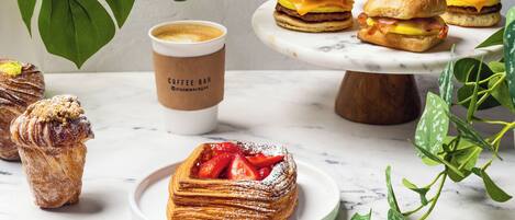 A variety of pastries and sandwiches on a marble table with a coffee cup and green plants in the background.