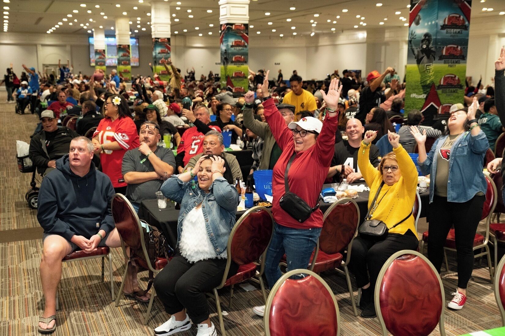 Un evento interior concurrido, con gente sentada y de pie, algunas alzando las manos, en una sala con sillas y mesas rojas.