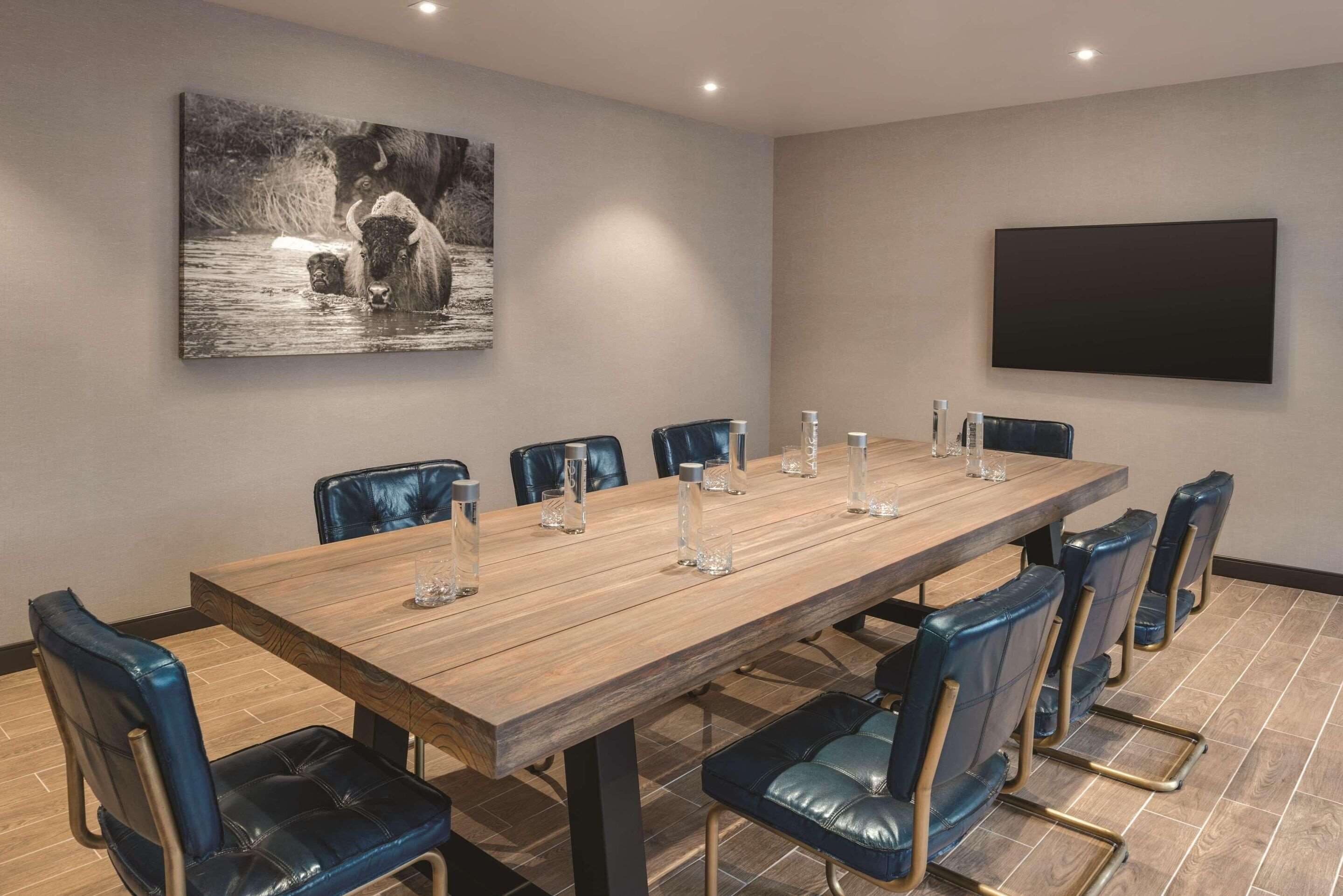 A conference room with a long wooden table, blue leather chairs, and a mounted flat-screen TV.
