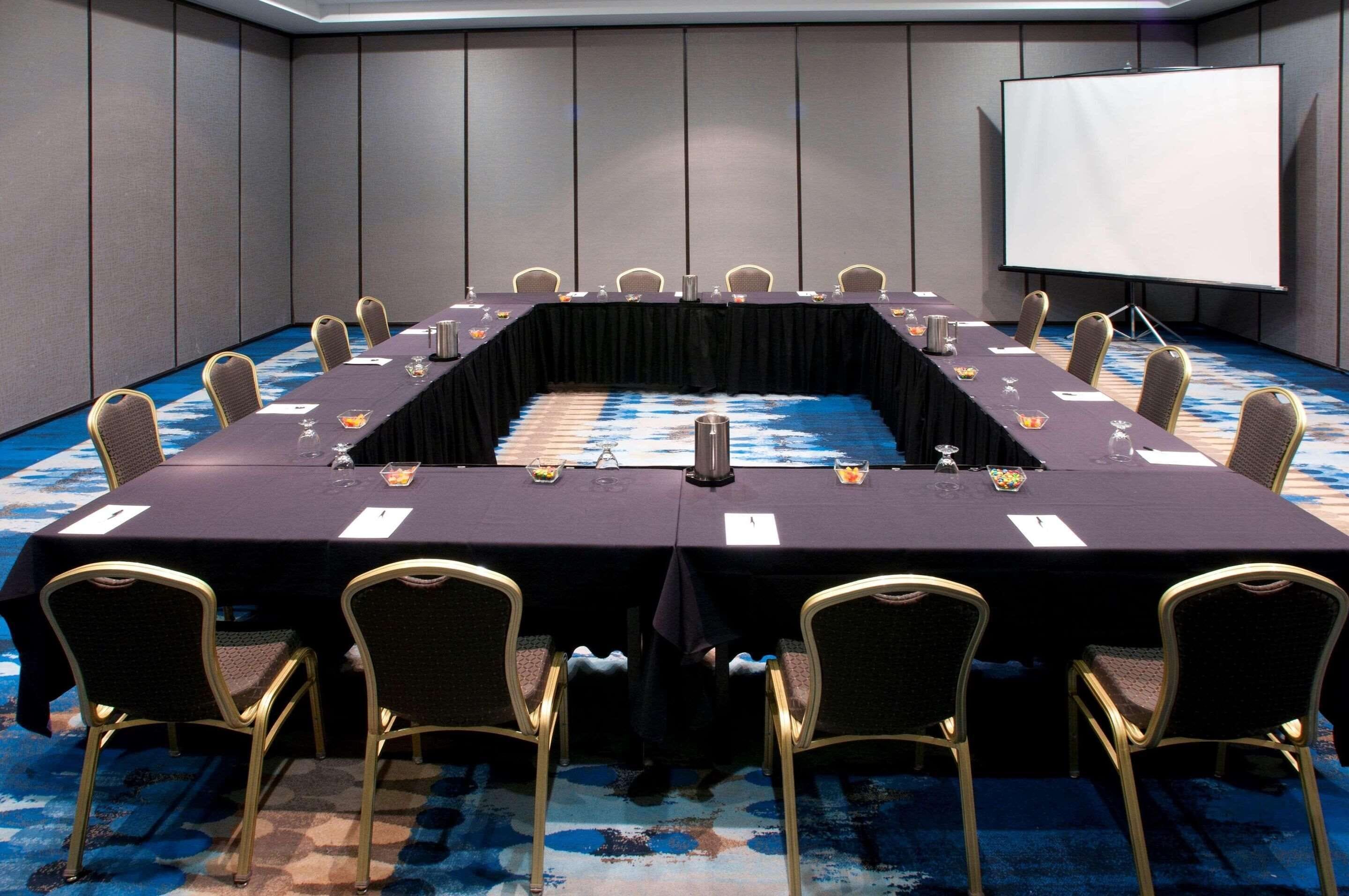 Un salón de conferencias con una larga mesa, sillas, una pantalla de proyector y suelo alfombrado.