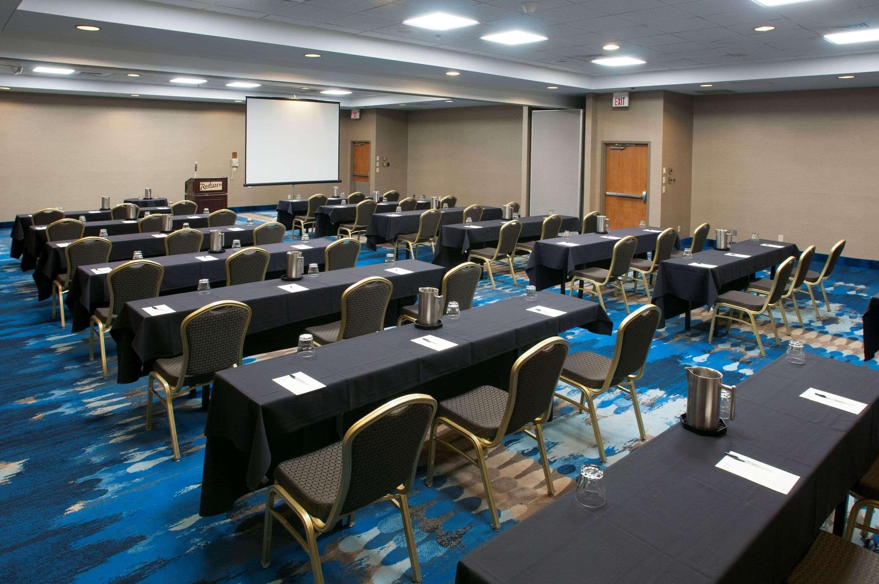 A conference room with rows of tables and chairs, a projector screen, and an exit door.