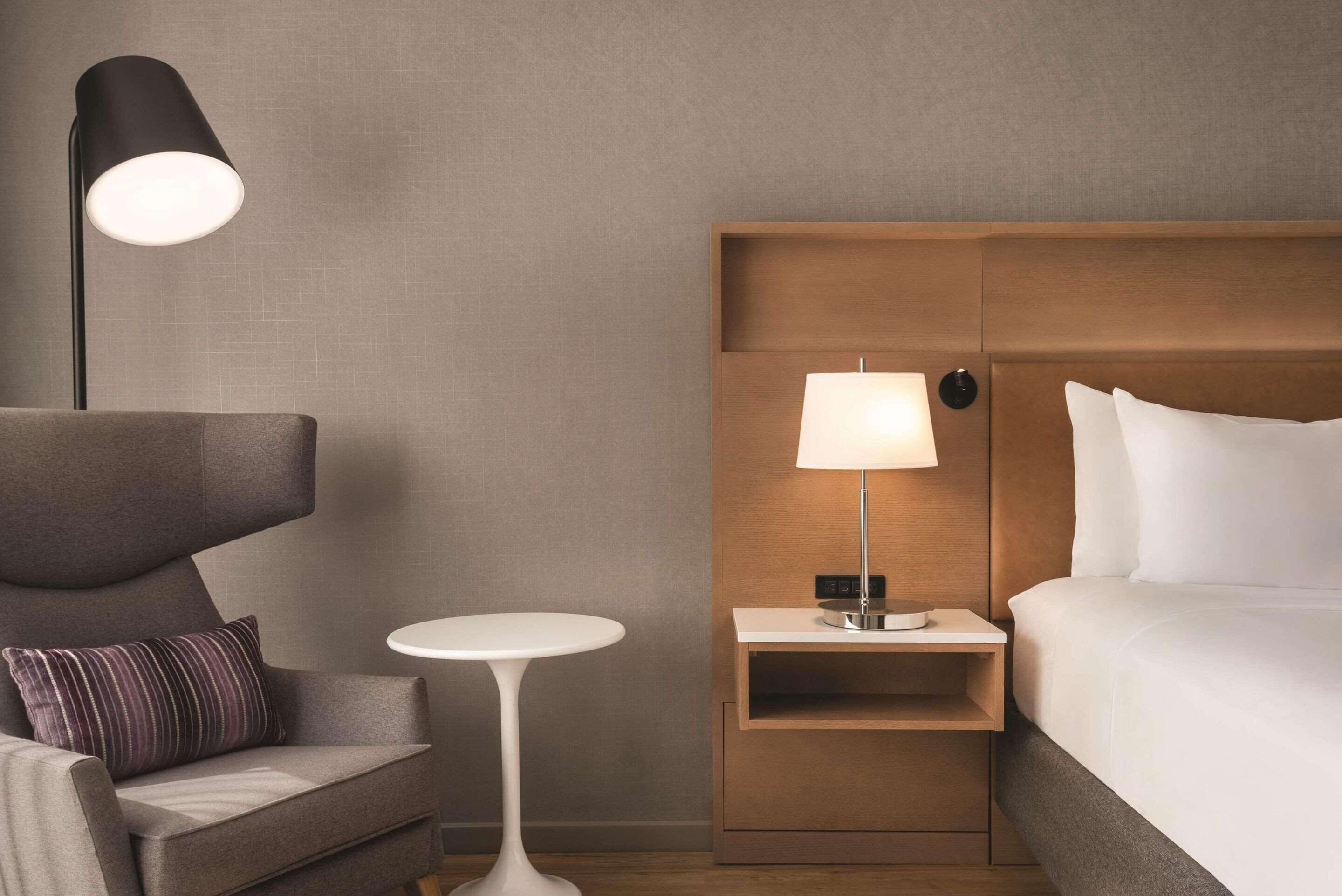 A hotel room with a modern design, featuring a bed, a nightstand with a lamp, a small round table, and a sofa with a pillow.