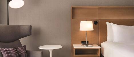 A hotel room with a modern design, featuring a bed, a nightstand with a lamp, a small round table, and a sofa with a pillow.
