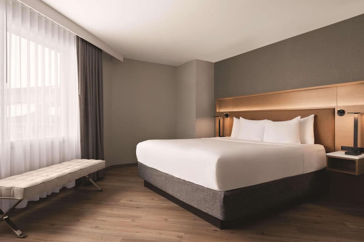 A modern hotel room with a large bed, bedside tables, a bench, and a view of the outdoors through sheer curtains.