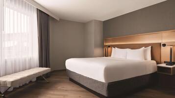 Habitación de hotel moderna con una cama grande, mesitas de noche, un banco y vistas al exterior a través de cortinas translúcidas.
