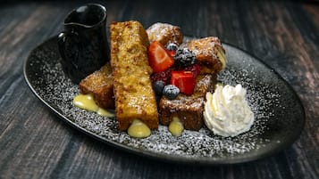 Una porción de tostadas francesas con fresas, arándanos y crema batida, sobre un plato oscuro espolvoreado con azúcar glas.