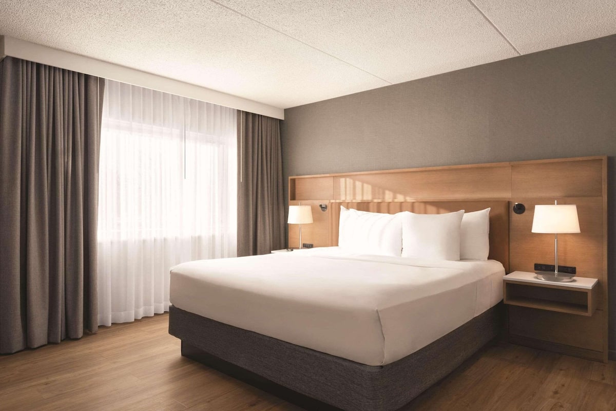 A hotel room with a large bed, two bedside tables with lamps, a wooden headboard, and a view of the outdoors through sheer curtains.