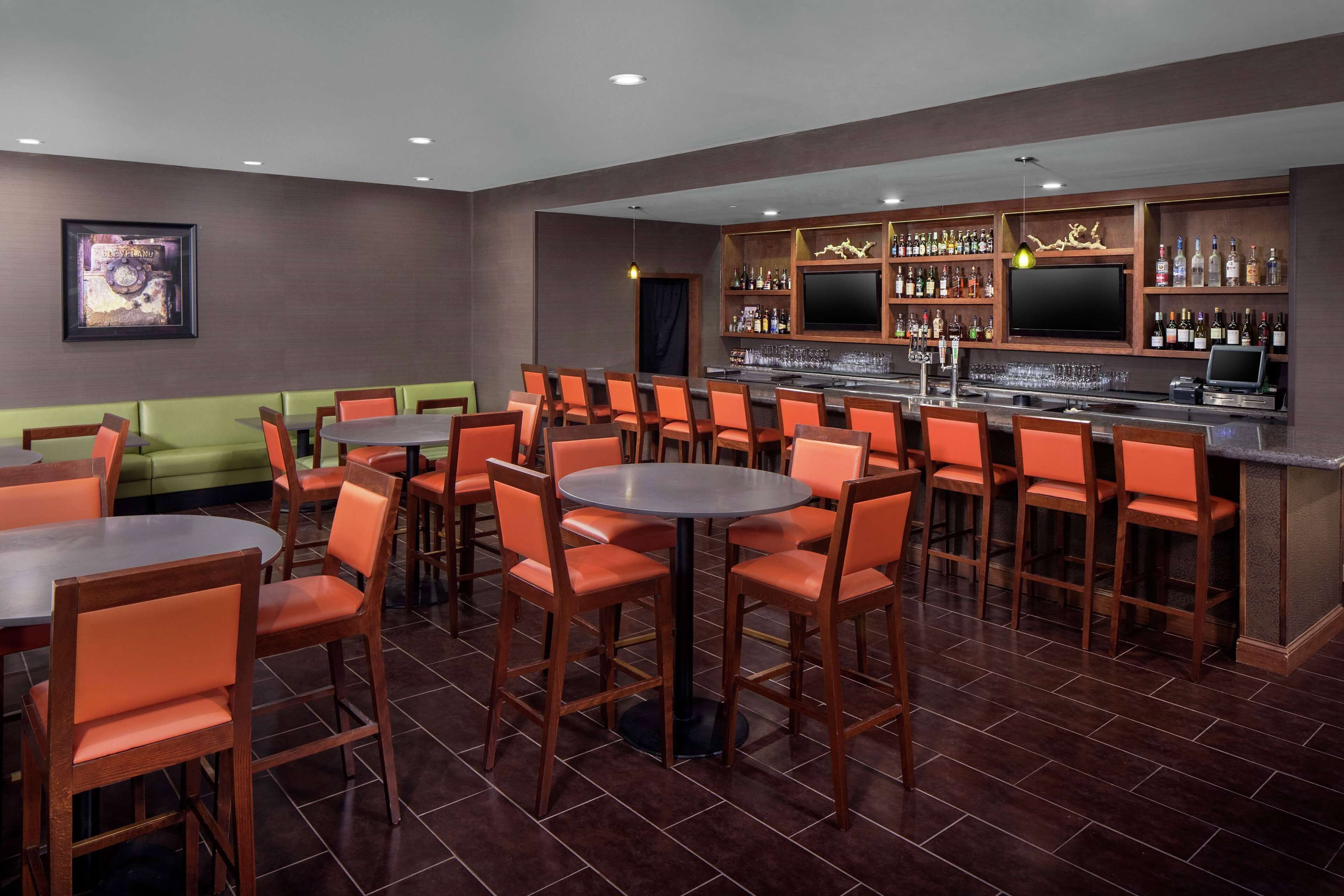 A bar area with multiple seating options including round tables and booths, a bar counter with a TV, and a variety of bottles and glasses on shelves.