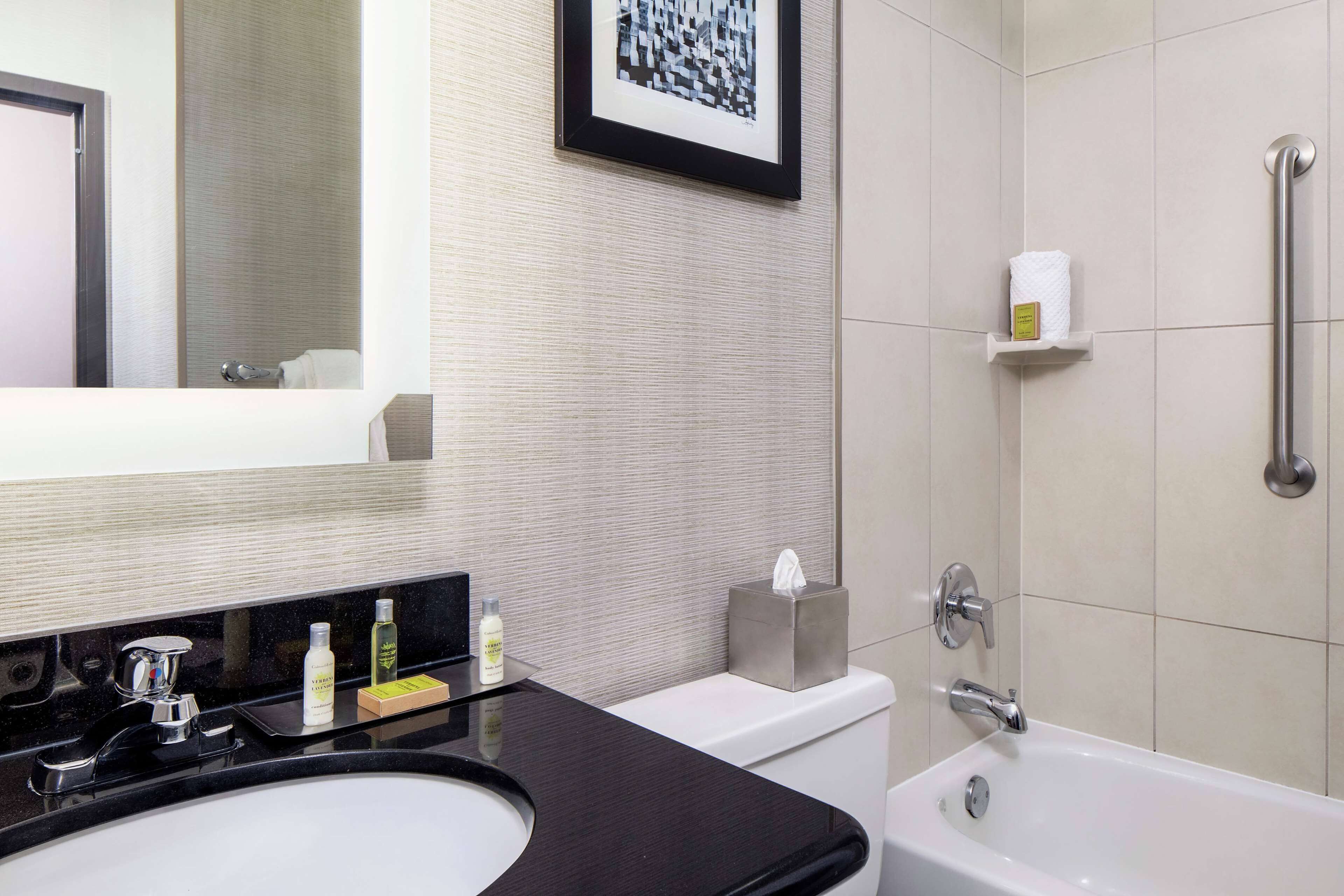 A modern bathroom with a black sink, white toilet, and a bathtub. There is a framed picture on the wall, a towel rack, and a shelf with toiletries.