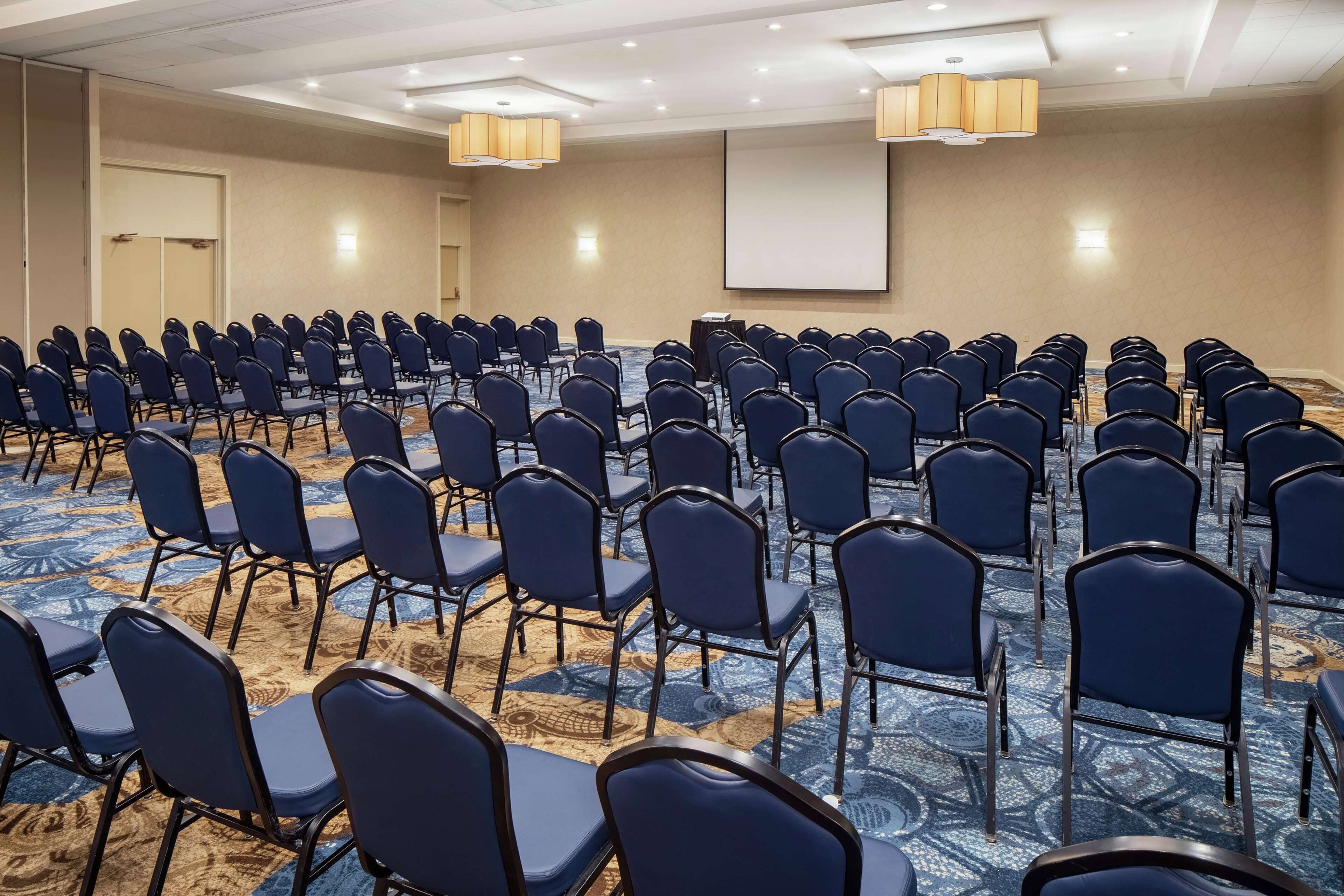 Una sala de conferències amb files de cadires blaves, un paviment amb moqueta i una pantalla de projector a la paret.
