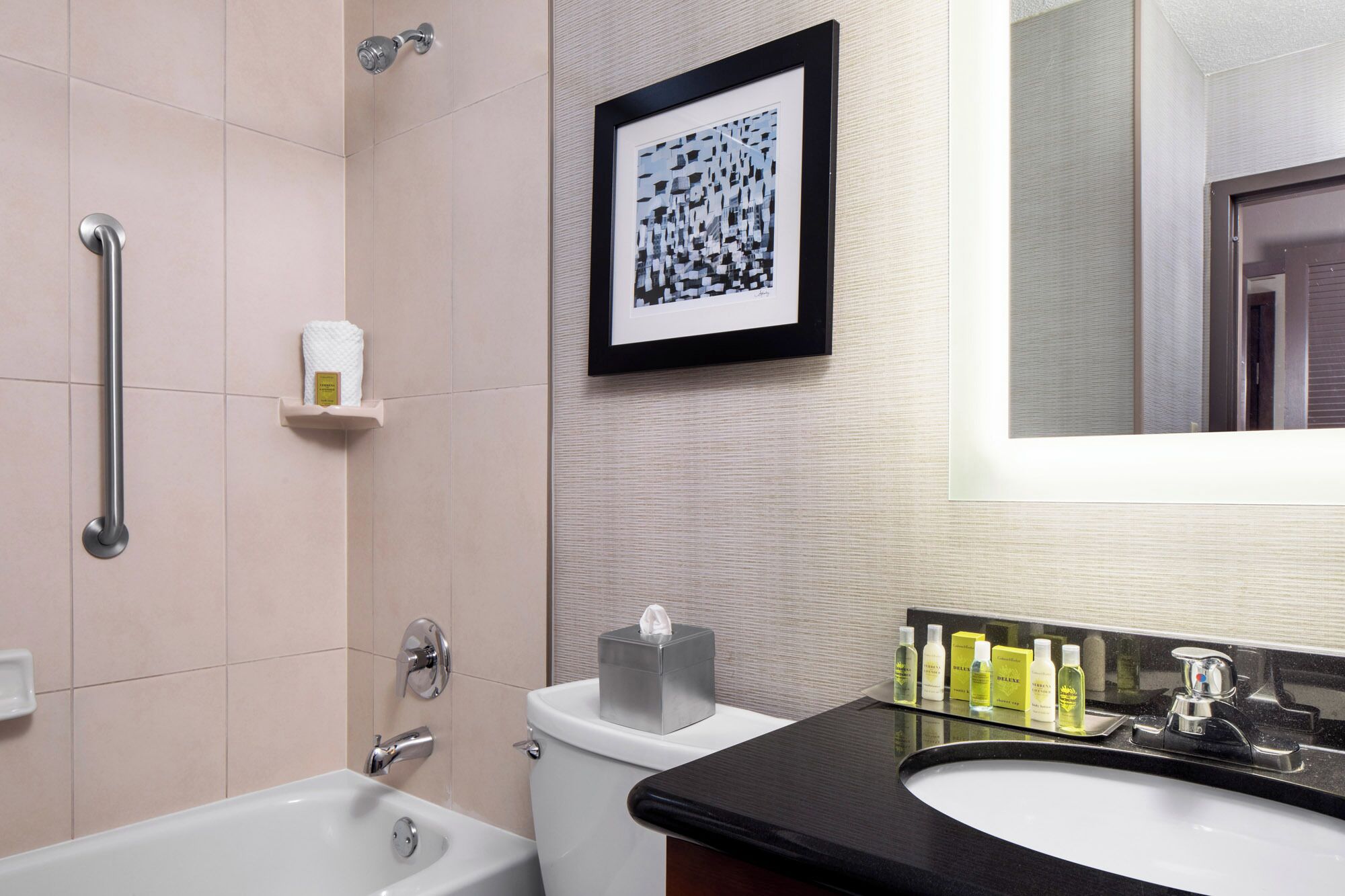 Une salle de bain moderne avec une baignoire, des toilettes et un lavabo. Un tableau encadré est accroché au mur, et des articles de toilette sont posés sur le comptoir du lavabo.