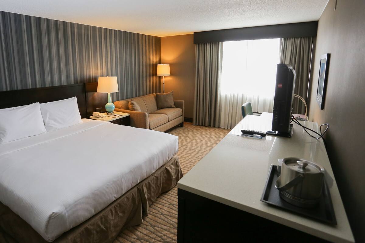 Habitación de hotel con una cama grande, un sofá, un escritorio con televisión y hervidor, y una ventana con cortinas.