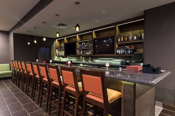 A bar area with a long counter, multiple chairs, and shelves stocked with bottles. - Wyndham Garden Westlake (Westlake)