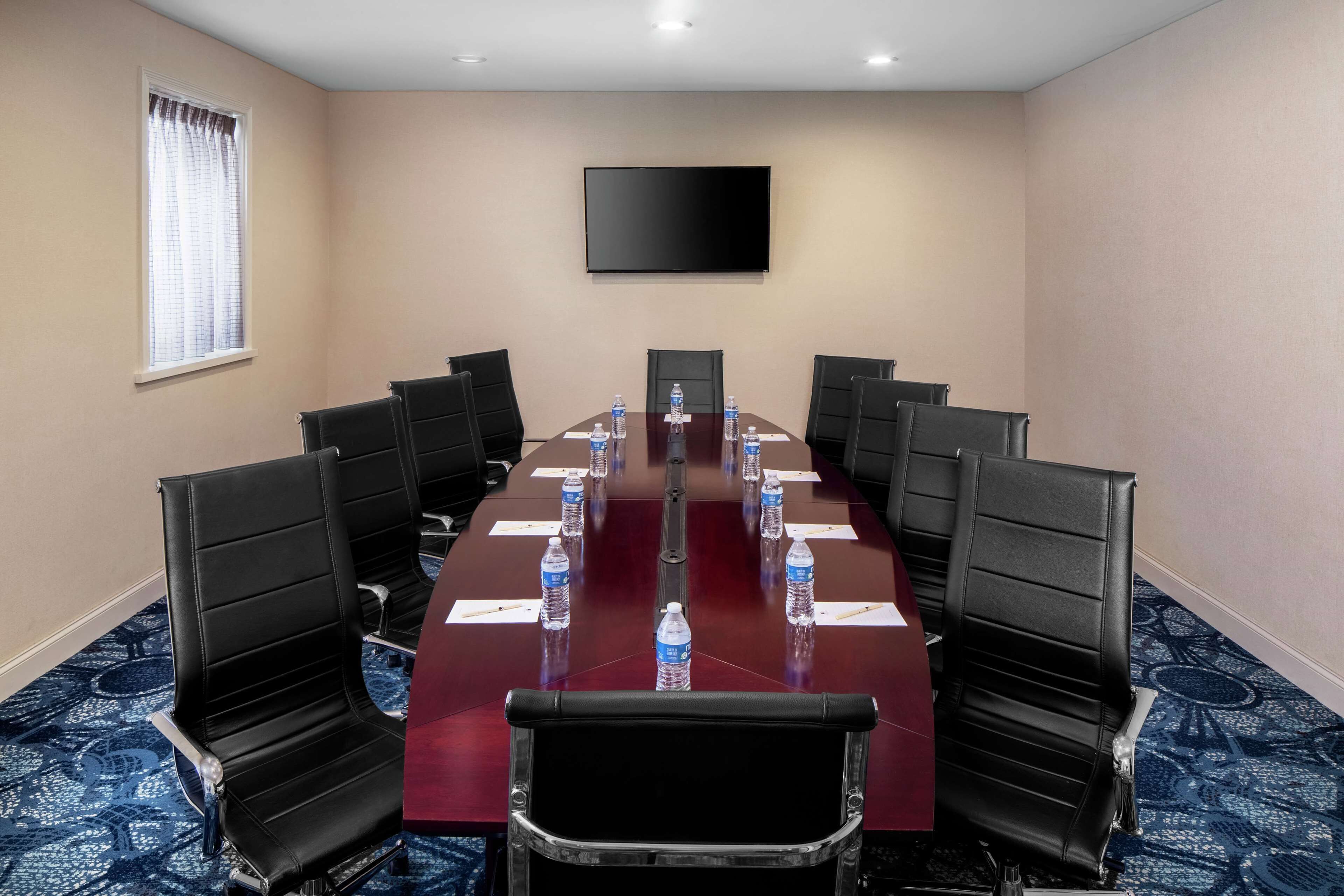Un salón de conferencias con una larga mesa, sillas negras, botellas de agua y un televisor de pantalla plana en la pared.