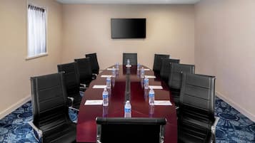 Un salón de conferencias con una larga mesa, sillas negras, botellas de agua y un televisor de pantalla plana en la pared.
