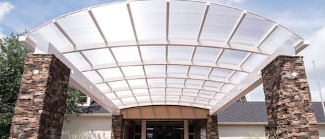A building with a large glass canopy entrance, stone pillars, and a paved walkway leading to the doors.