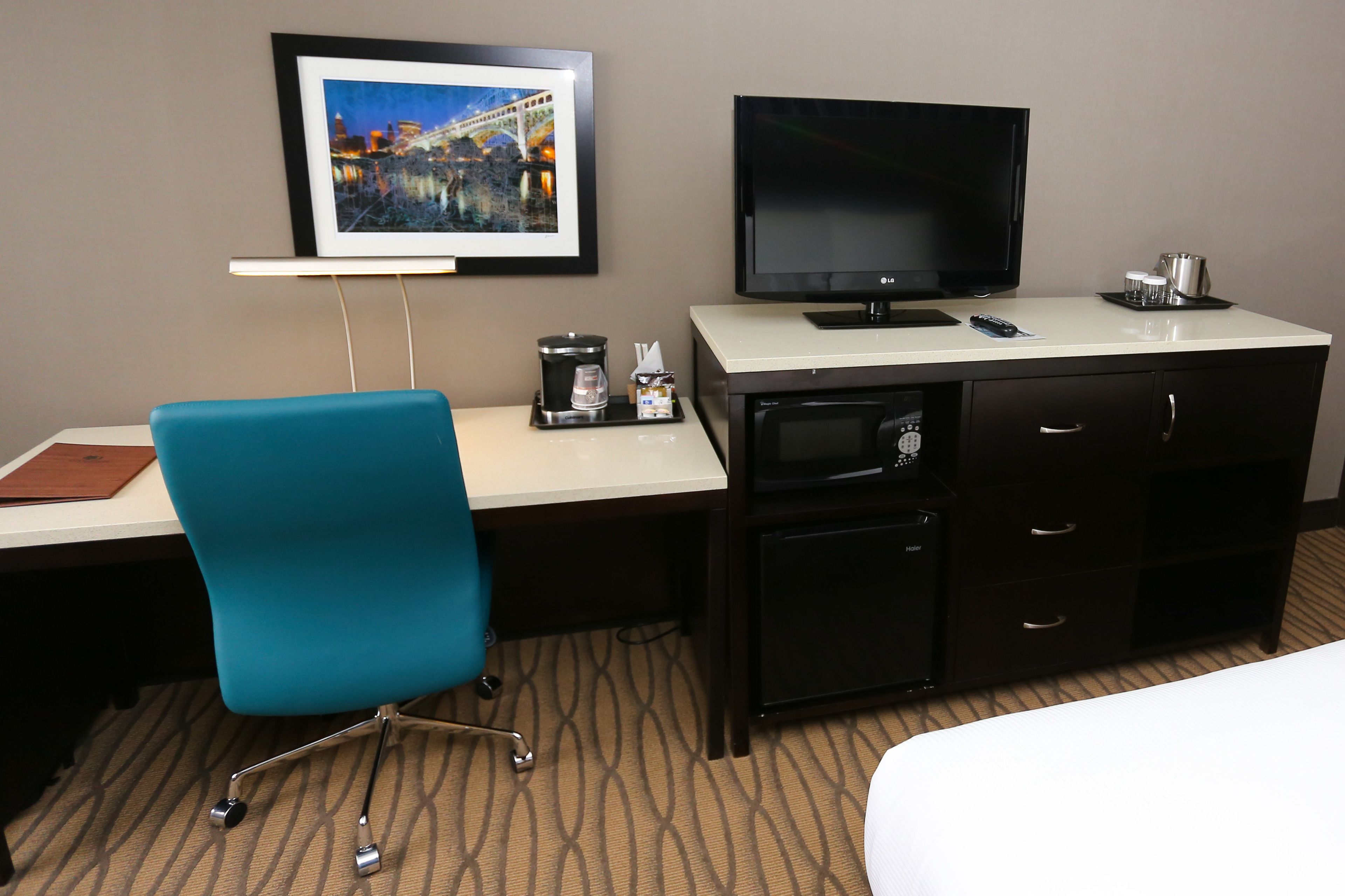 A hotel room with a bed, a desk with a computer, a TV, a coffee maker, and a framed picture on the wall.