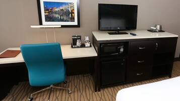 A hotel room with a bed, a desk with a computer, a TV, a coffee maker, and a framed picture on the wall.
