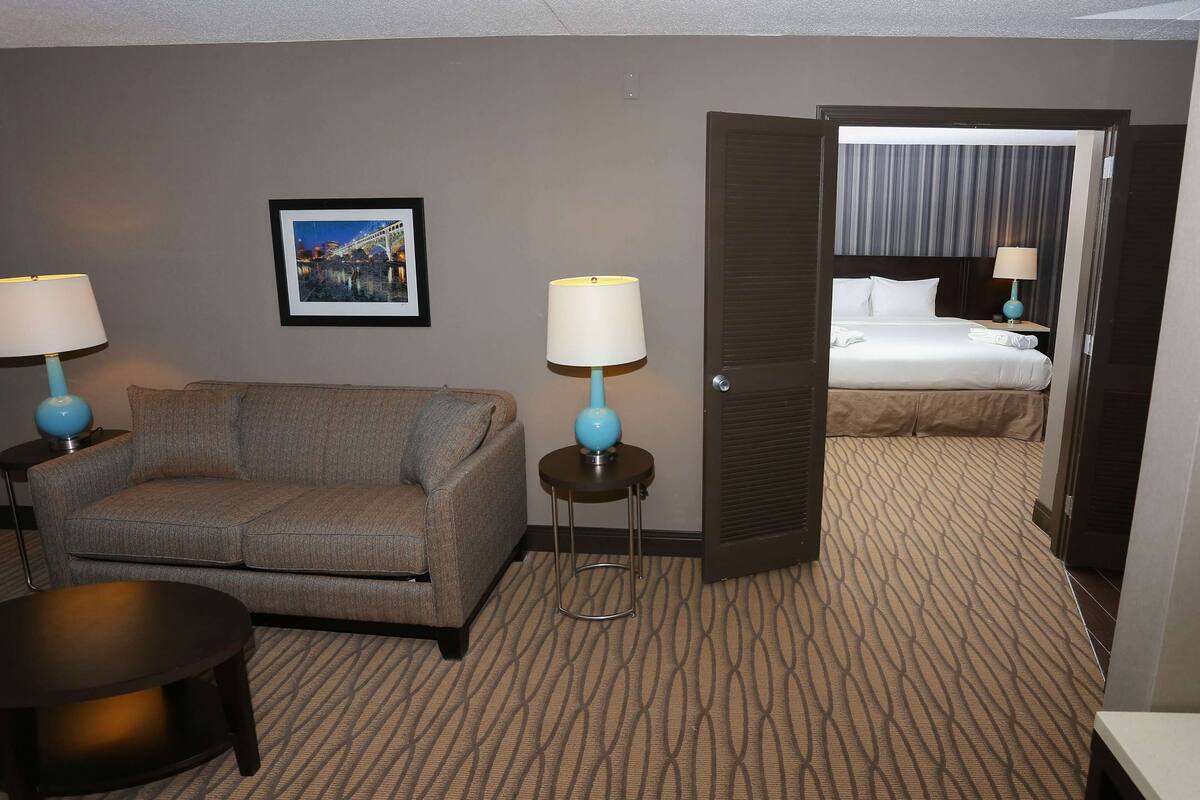 Habitación de hotel con sofá, dos mesitas de noche con lámparas, cama con cabecero y alfombra estampada.