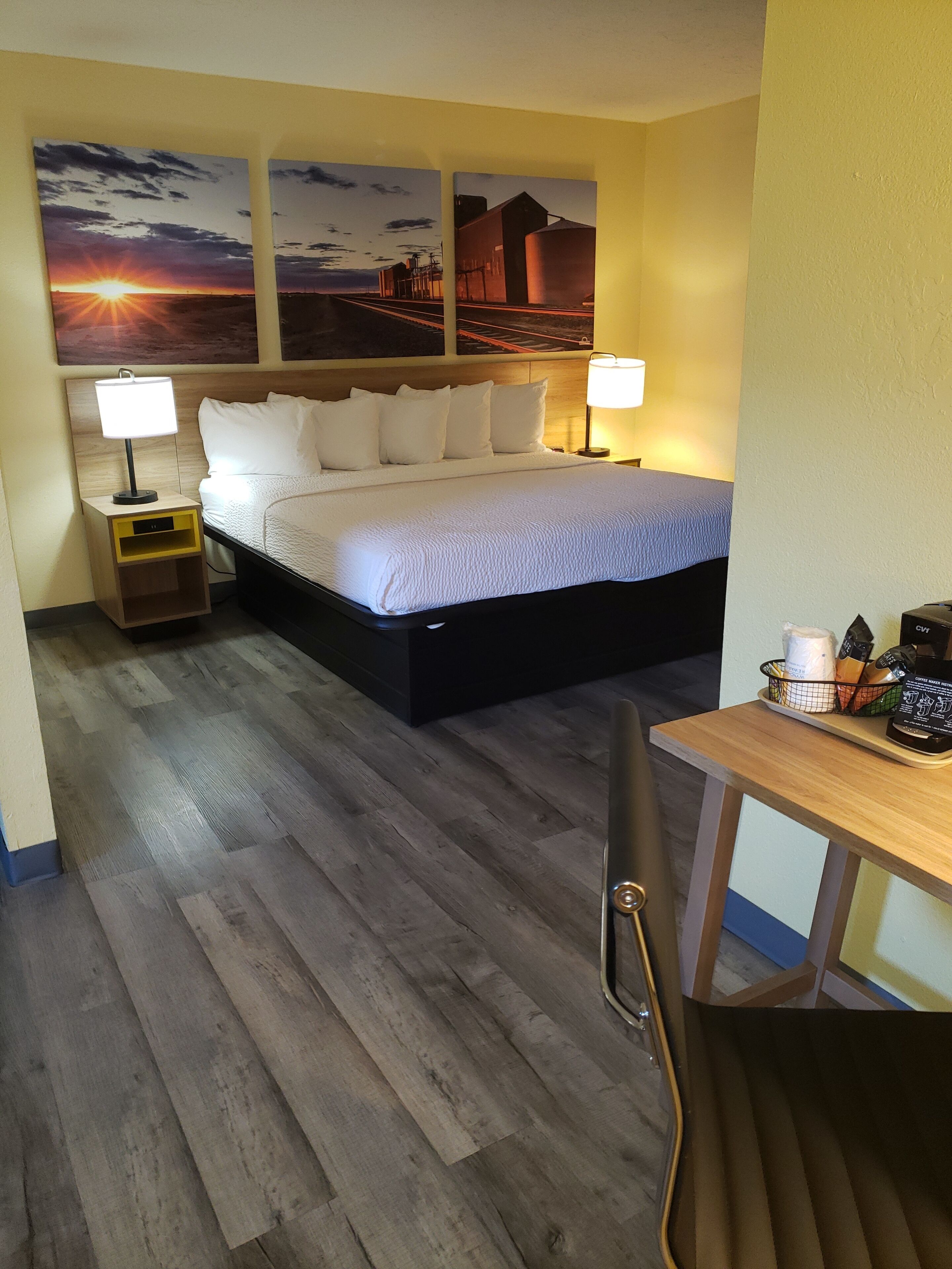 A hotel room with a bed, two bedside lamps, a nightstand, a desk with a chair, and two wall-mounted paintings.