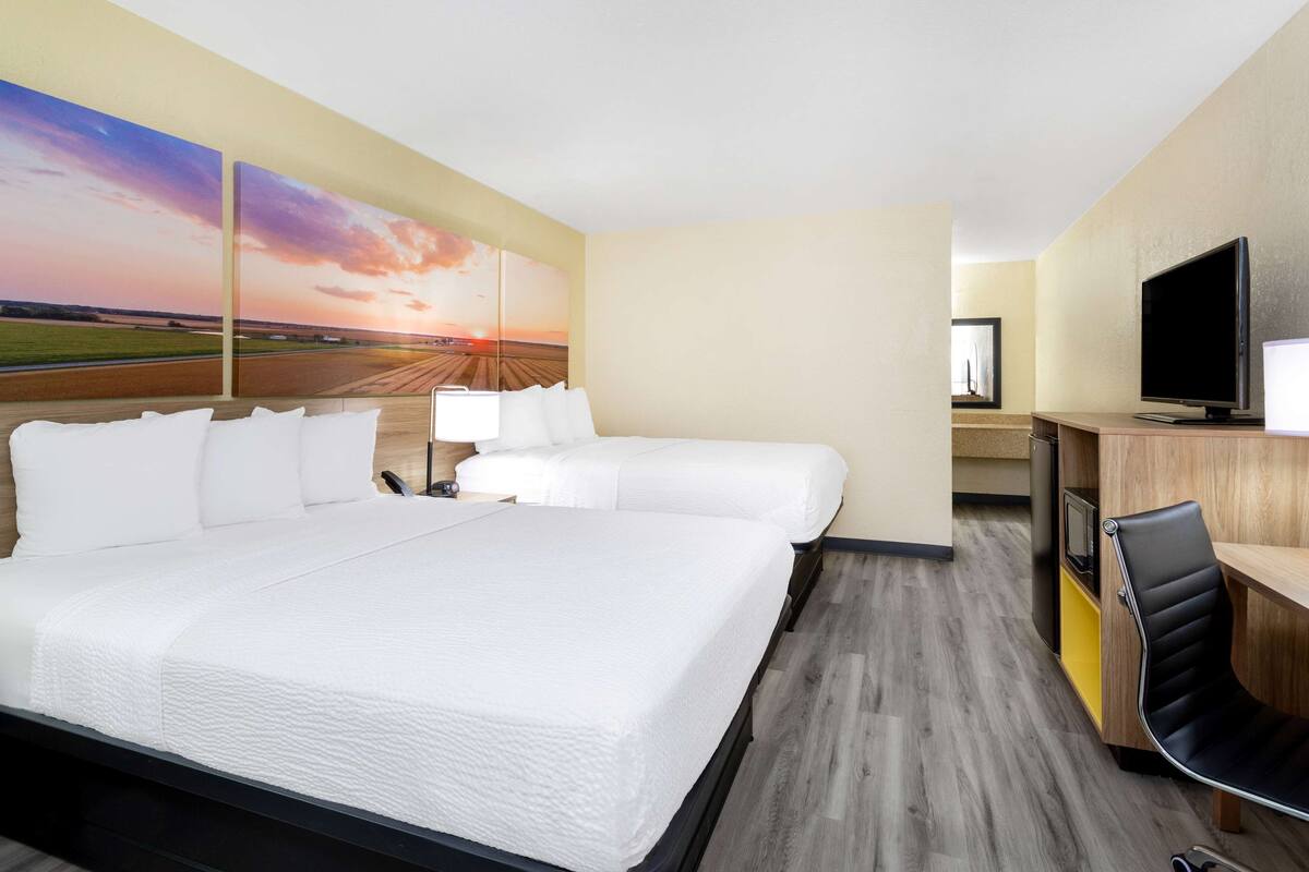 Quarto de hotel com uma cama grande, duas camas menores, uma escrivaninha com televisão e um quadro de pôr do sol na parede.