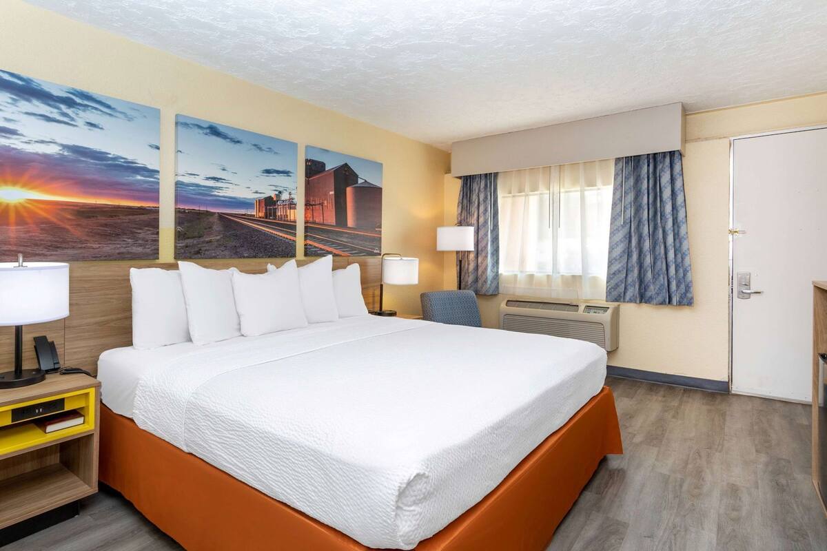 Quarto de hotel com uma cama grande, uma mesa com luminária, uma cadeira, uma janela com cortinas e obras de arte na parede.