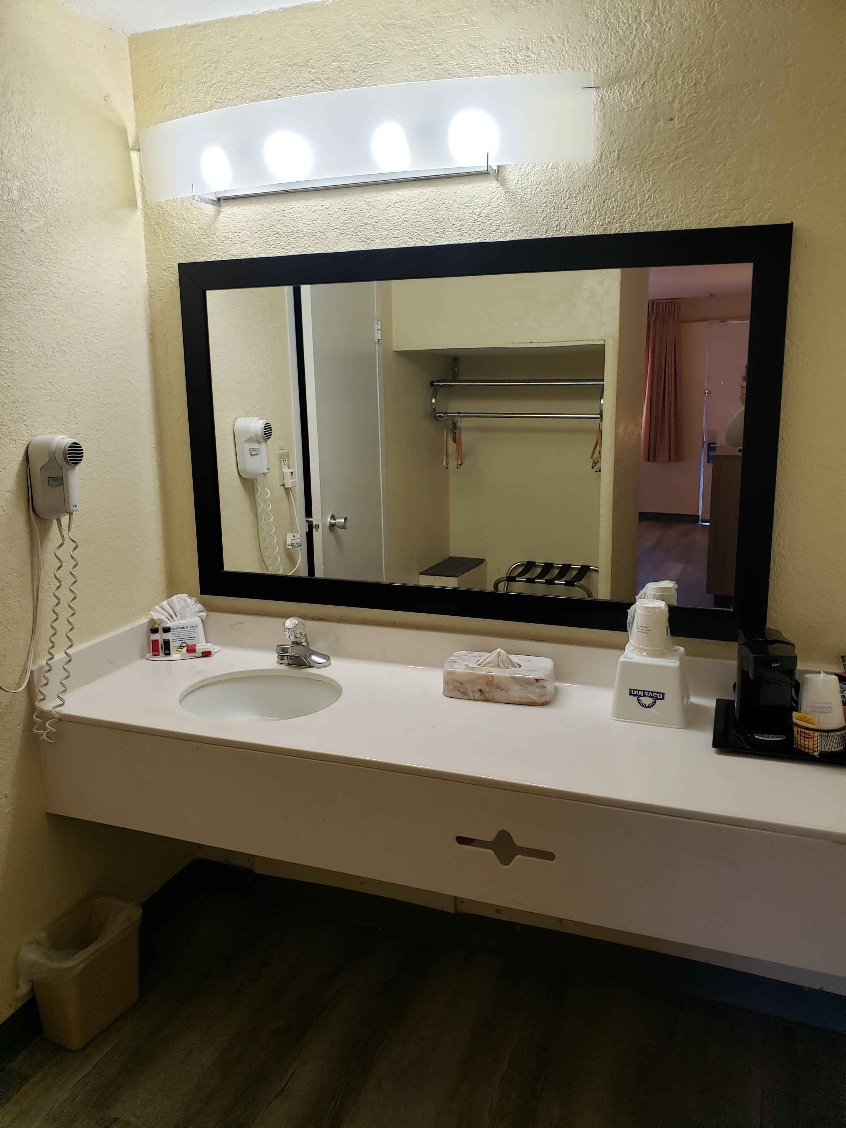 A hotel bathroom with a large mirror, a sink, a hairdryer, a towel, a coffee maker, and a trash bin.
