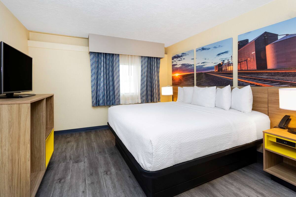 Quarto de hotel com uma cama grande, uma TV, uma mesa e uma pintura de um pôr do sol sobre trilhos de trem.
