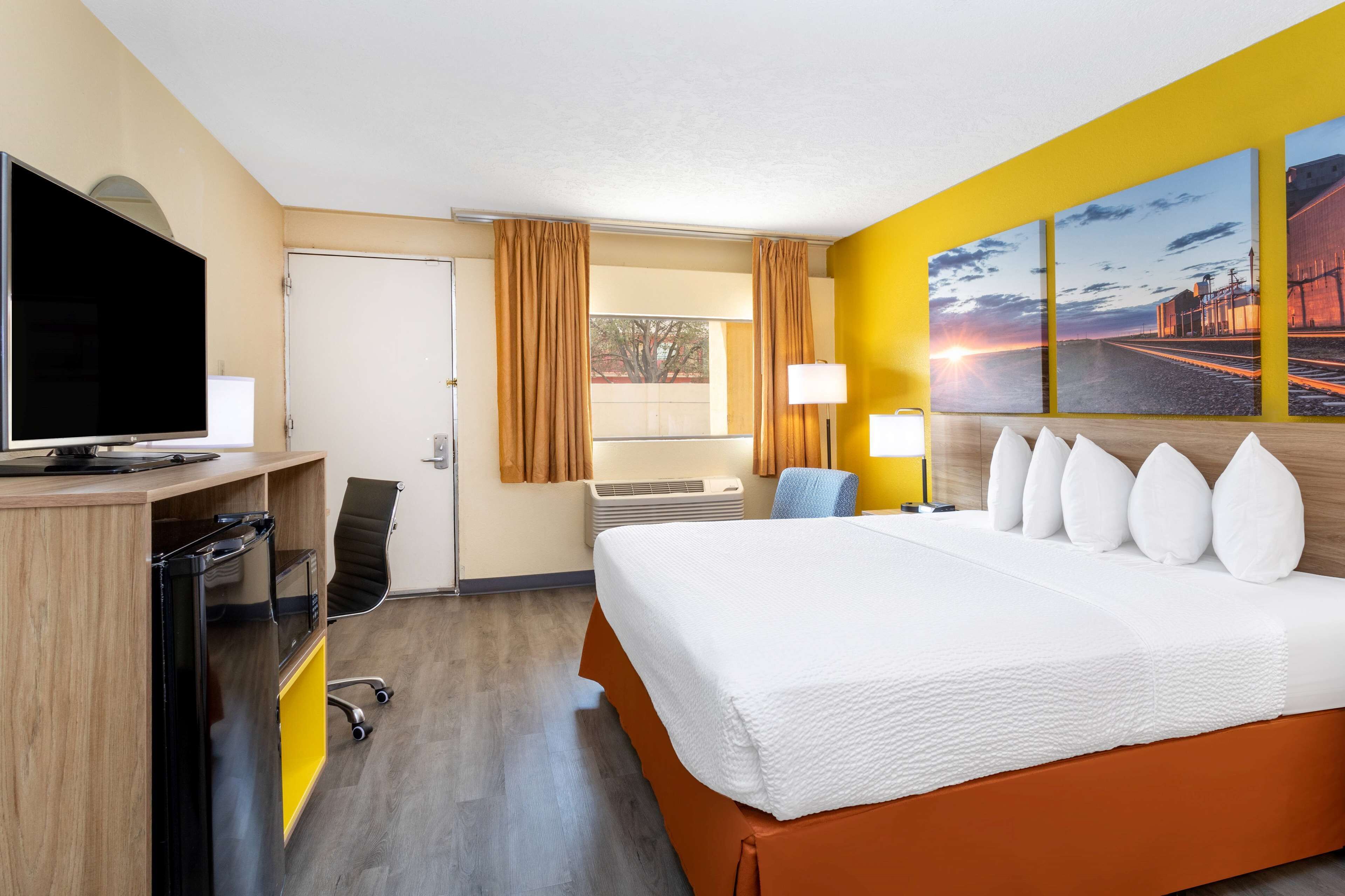 A hotel room with a bed, a desk with a TV, a chair, a window with curtains, an air conditioning unit, and artwork on the wall.