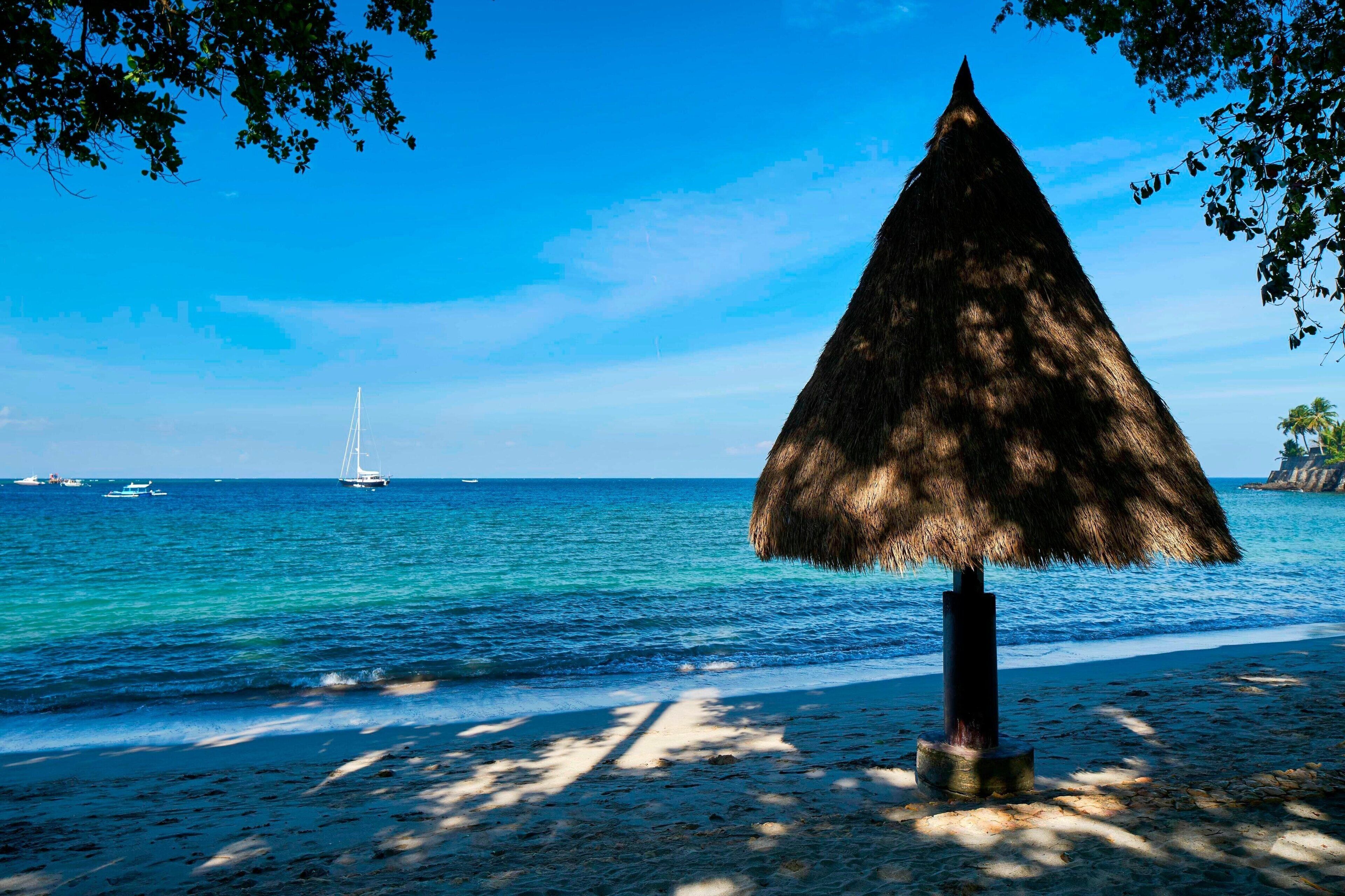 Une plage avec un parasol en chaume, une eau d’un bleu limpide et des bateaux à l’horizon.