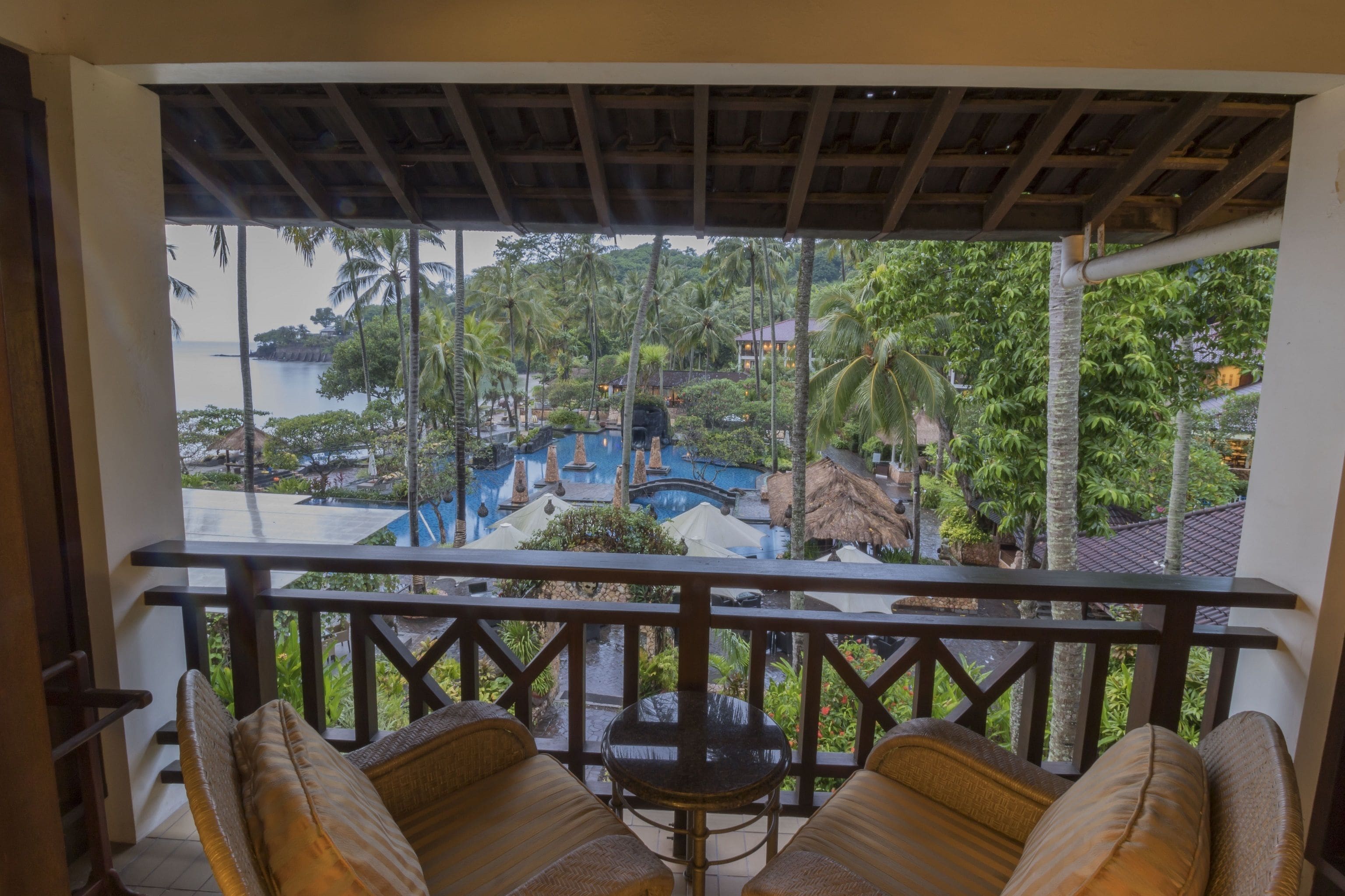 Varanda com móveis de vime e vista para um resort tropical com piscina e palmeiras.