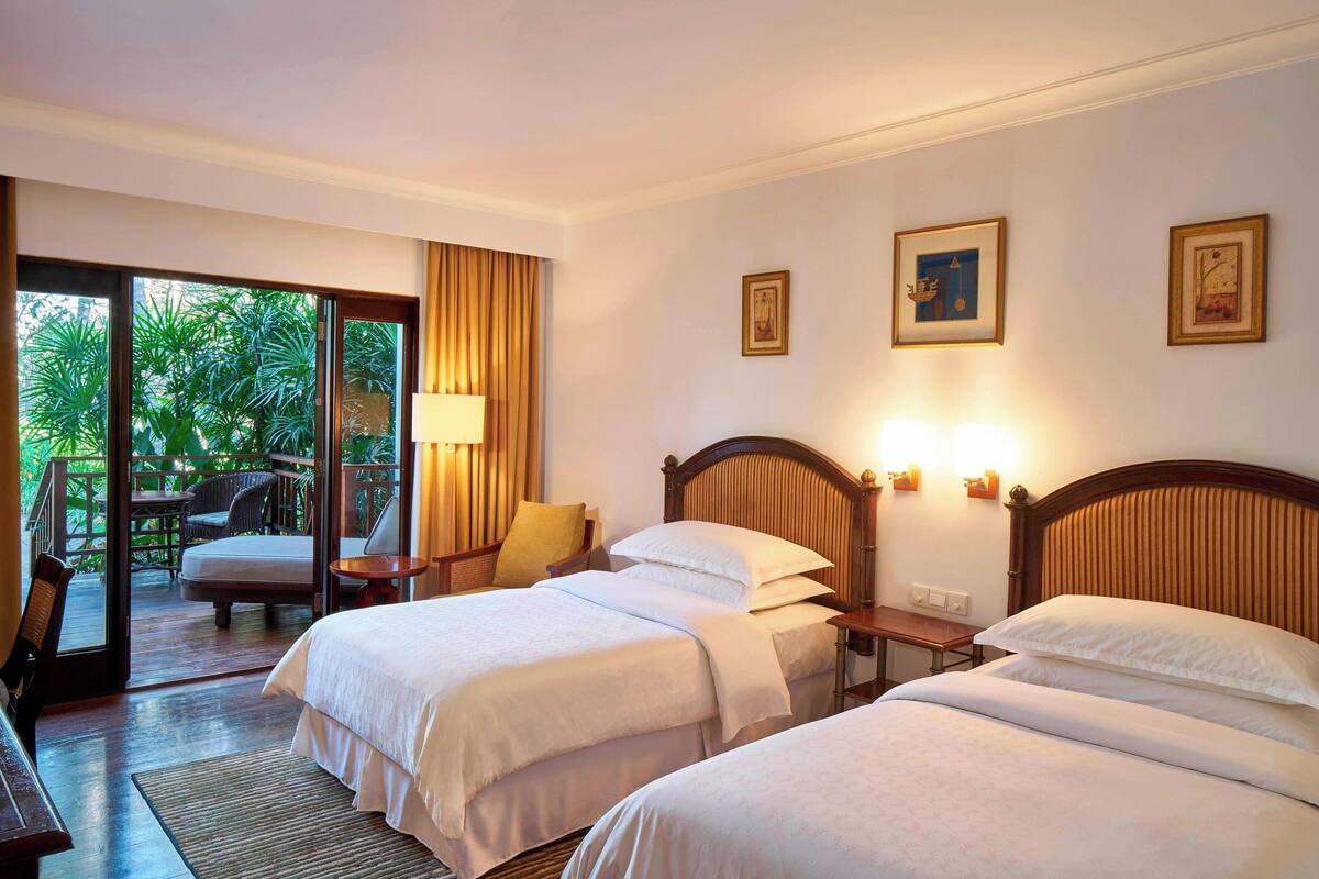 A hotel room with two beds, a wooden headboard, a nightstand, a desk, and a chair. There is a balcony with a view of greenery.