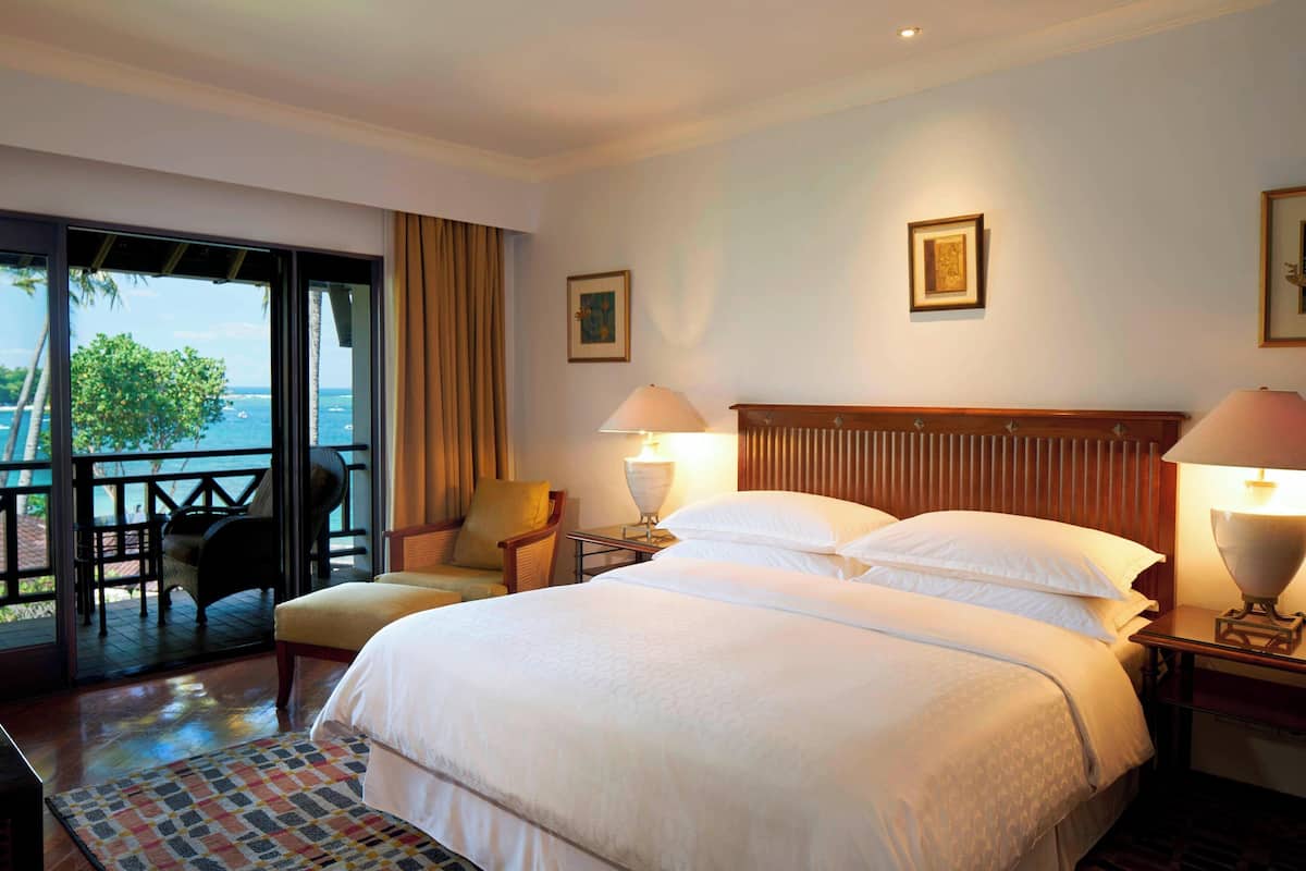 A hotel room with a large bed, two bedside lamps, a chair, and a view of the sea through a sliding door.