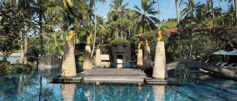 A clear blue pool with stone pillars and a stone bridge, surrounded by palm trees.