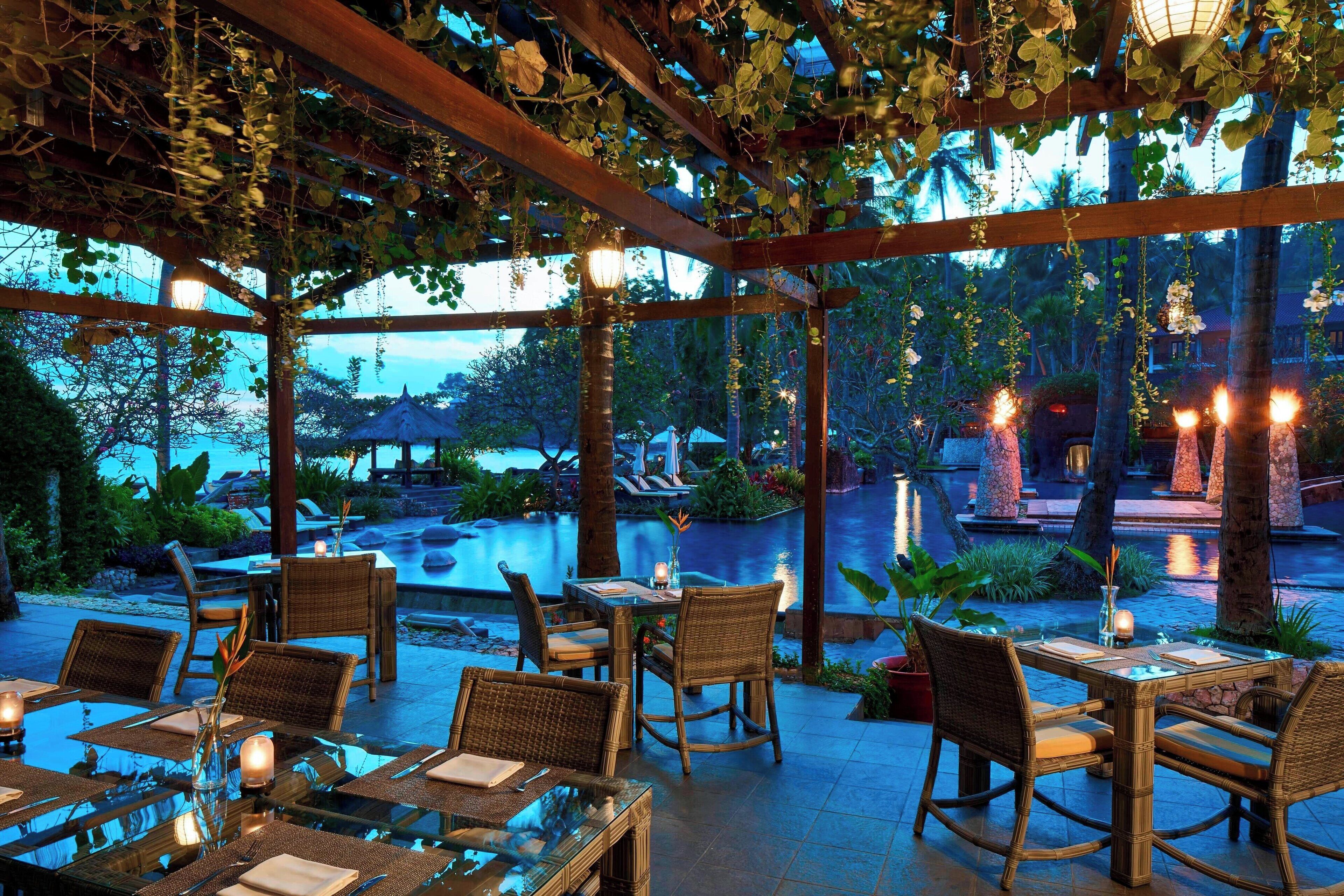 An outdoor dining area with wicker furniture and a view of a pool and tropical plants.