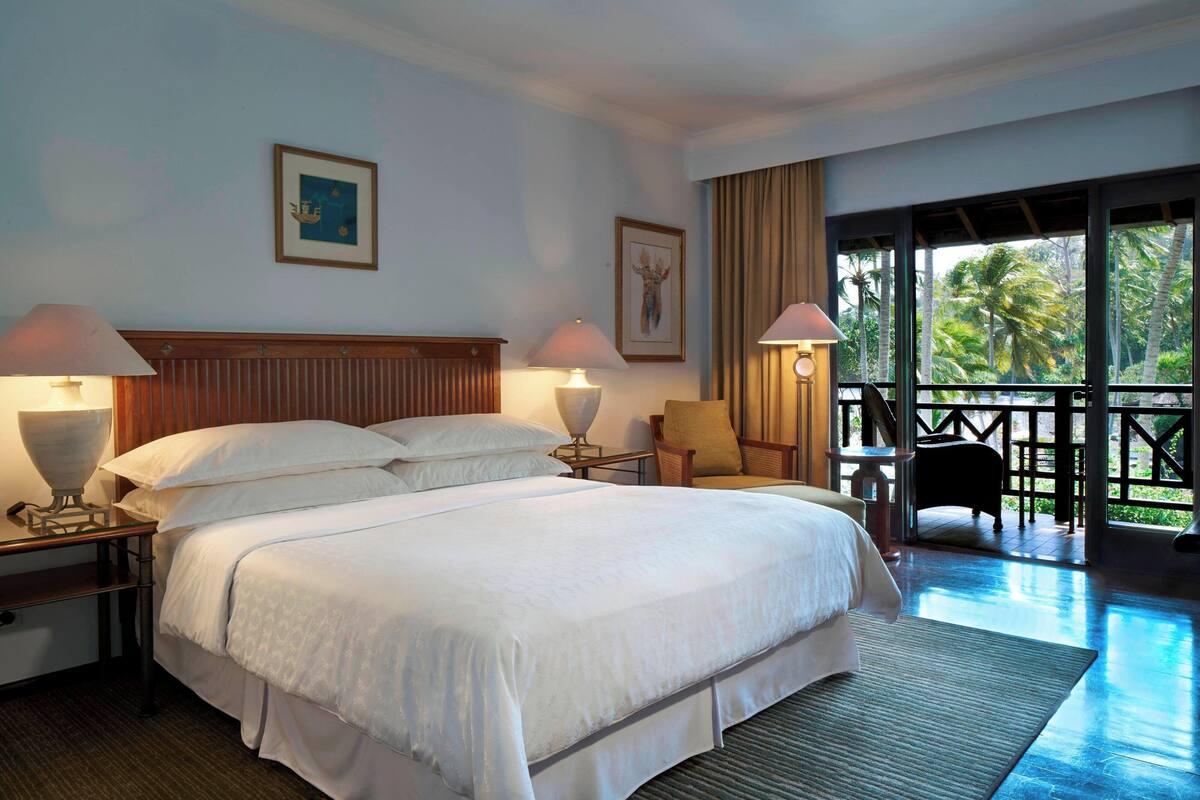 A hotel room with a large bed, two bedside tables with lamps, a chair, and a balcony with a view of palm trees.