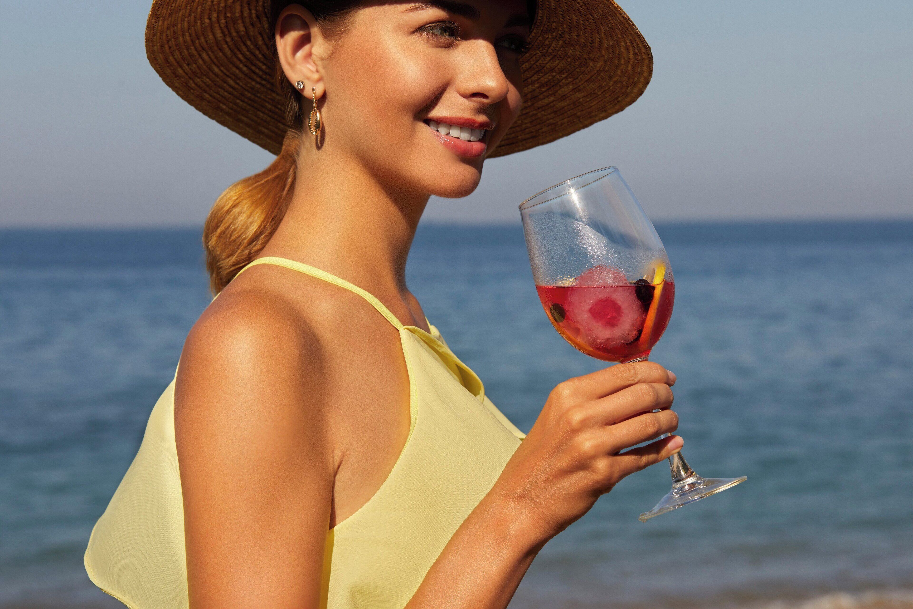 A person holding a glass of red drink with a straw, wearing a yellow top and a straw hat, with a body of water in the background.