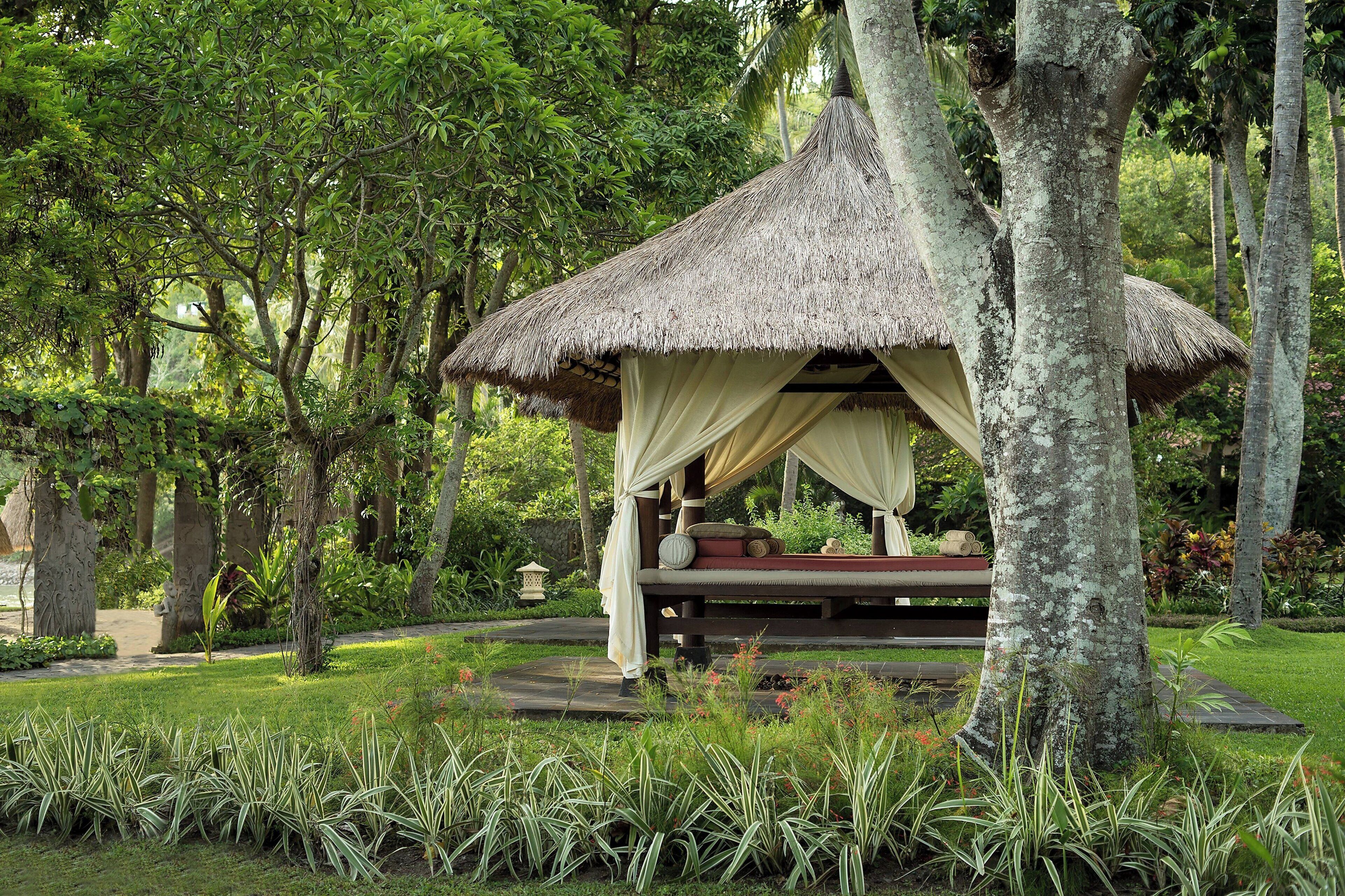 A thatched-roof gazebo with a bench and cushions, surrounded by lush greenery and trees.