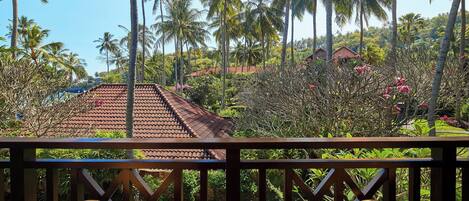A wooden deck with wicker furniture, overlooking a tropical landscape with palm trees and a red-roofed building.