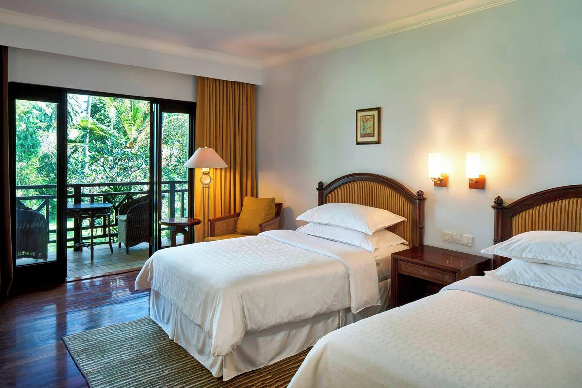 A hotel room with two beds, a balcony with a table and chairs, and a view of greenery.